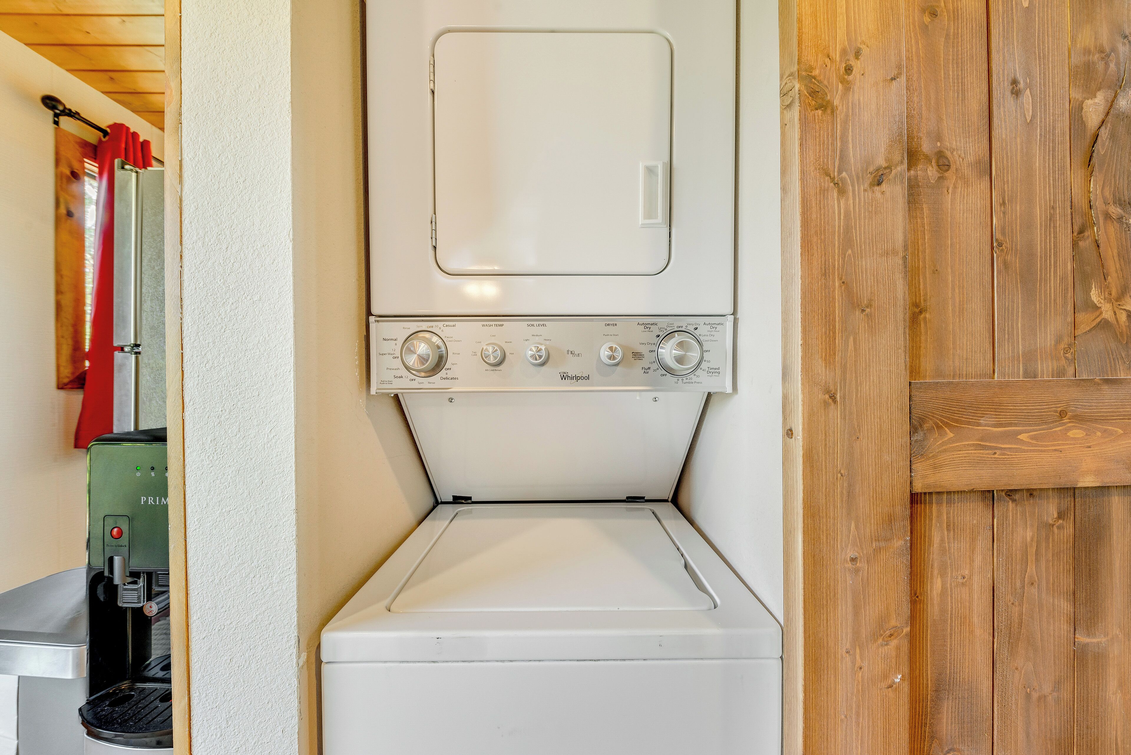 Una lavadora blanca y compacta con perillas de control y puerta frontal, ubicada en una habitación con paredes de madera y una cortina roja visible.