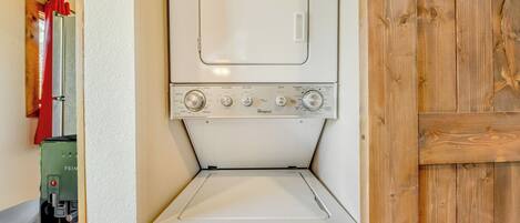 Una lavadora blanca y compacta con perillas de control y puerta frontal, ubicada en una habitación con paredes de madera y una cortina roja visible.