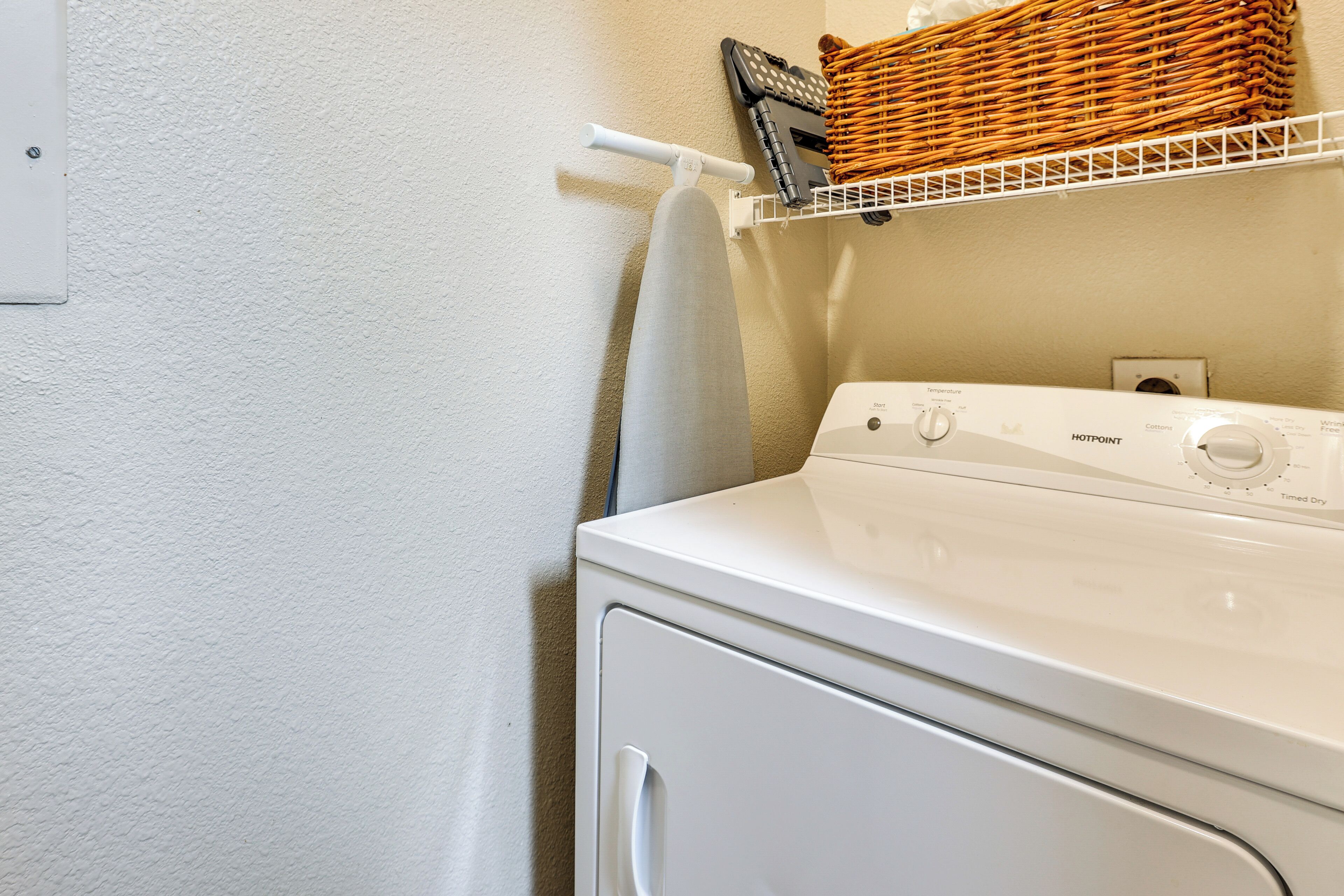 A front-loading washing machine with a control panel, a white wall, and a wicker basket on a shelf above.