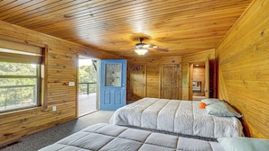 A room with wooden walls, a ceiling fan, two beds, a blue door, and a window. - Lake Eufaula Cabin w/ Hot Tub & Large Deck (Stigler)