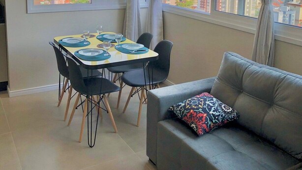 A modern living room with a dining area, large windows with city views, and a grey sofa with a patterned cushion. - Dallas Apartamentos (Bento Gonçalves)