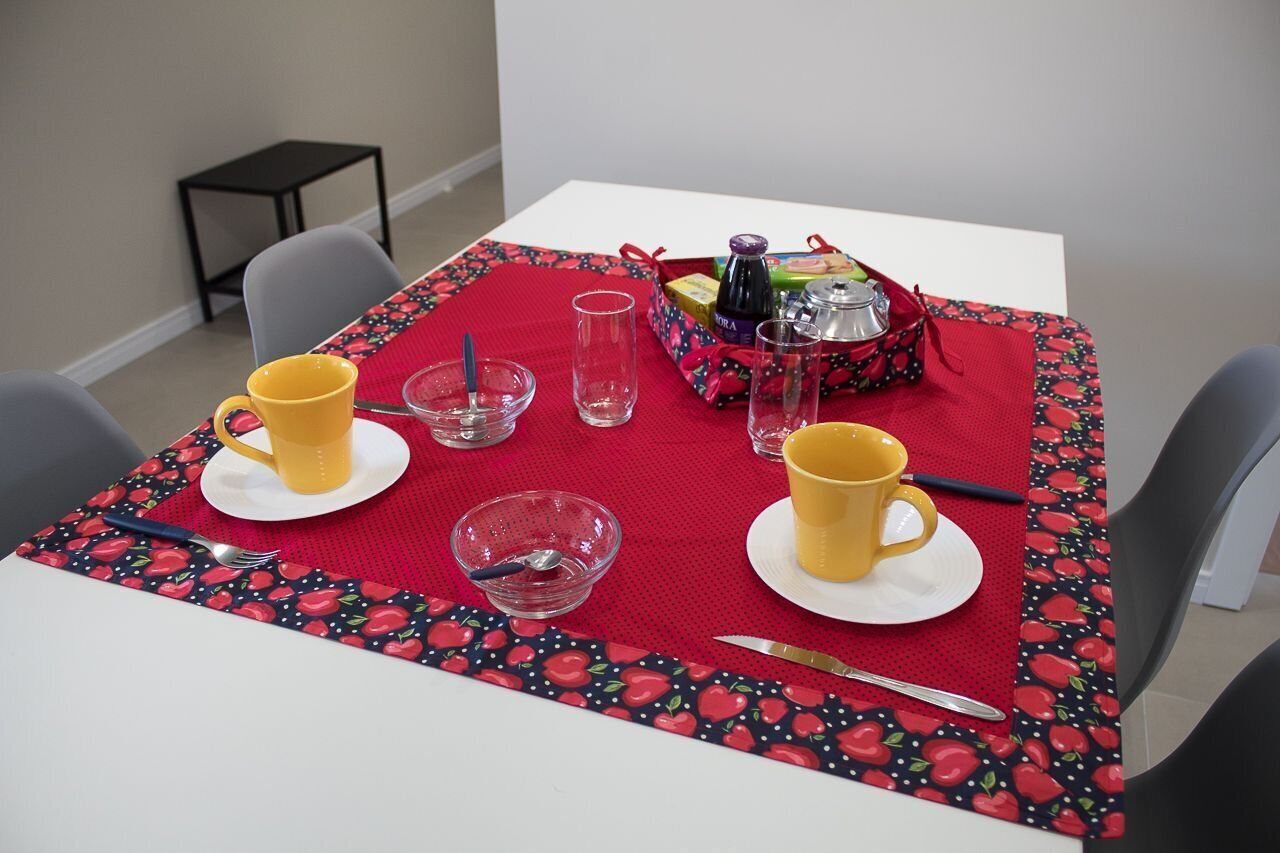 Une table à manger est dressée avec des tasses jaunes, des verres transparents, un bol et un couteau. Une nappe rouge à motifs de fruits recouvre la table, et une petite table noire se trouve à l’arrière-plan.