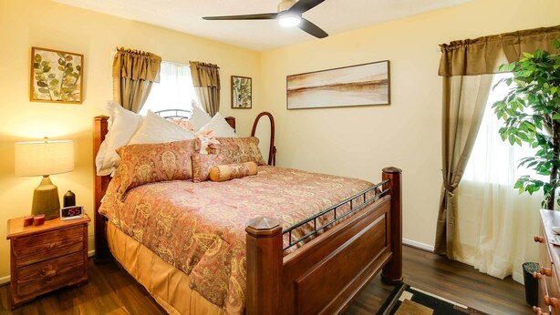 Un dormitorio con cama de madera, una mesita de noche con lámpara, ventilador de techo y un amplio ventanal con cortinas.
