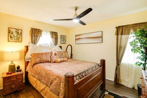 A bedroom with a wooden bed, a nightstand with a lamp, a ceiling fan, and a large window with curtains. - Lovely Compton Vacation Rental w/ Patio & Grills! (Compton)
