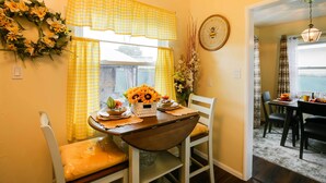 Un comedor con una mesa dispuesta para dos personas, cortinas amarillas, un reloj en la pared y un arreglo floral.