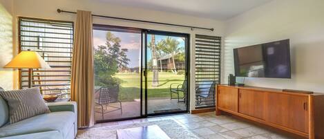 A living room with a sofa, a coffee table, a TV, and a sliding glass door leading to an outdoor area with a lawn and chairs.