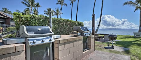 An outdoor barbecue area with a grill, a gas stove, and a charcoal grill, surrounded by palm trees and a view of the ocean.