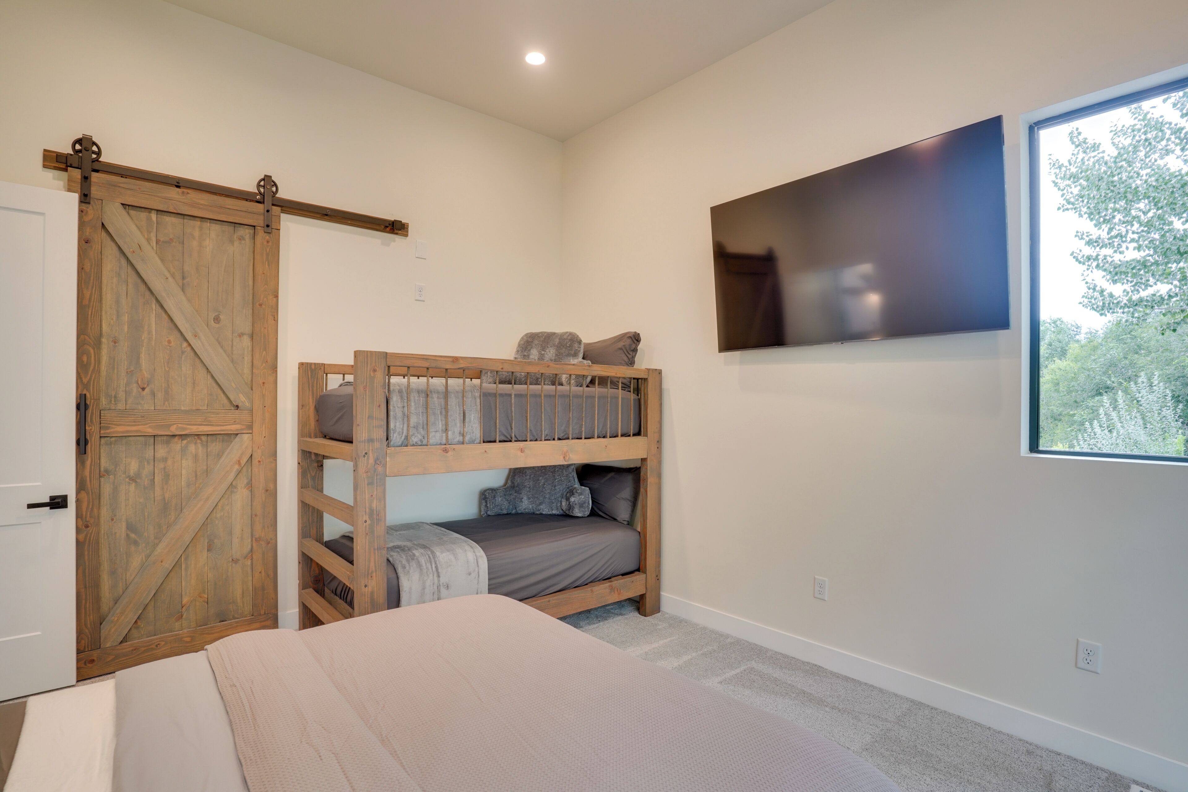 Quarto com beliche, porta corrediça em estilo rústico, televisão na parede e janela com vista para árvores.