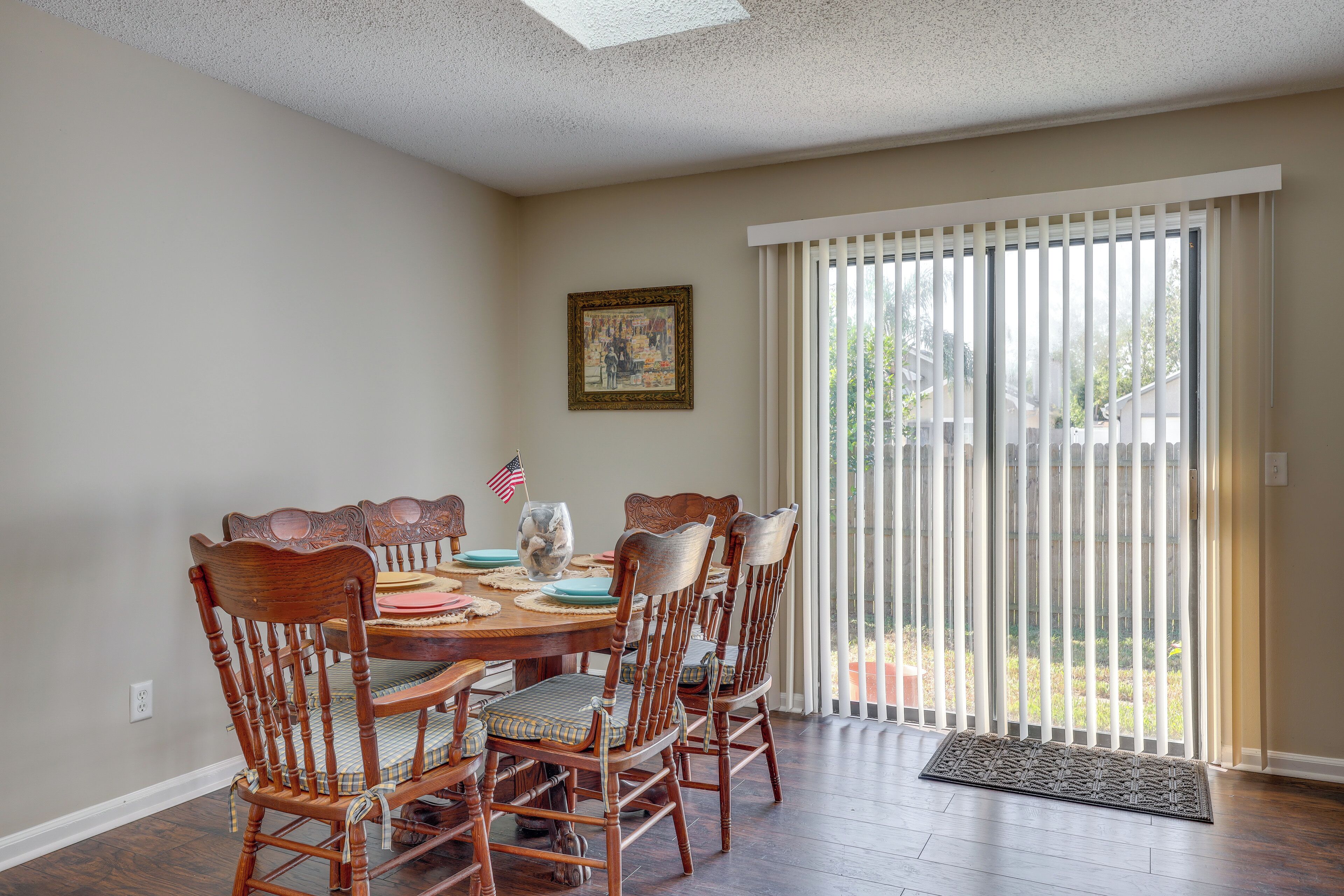 A dining area with a wooden table and chairs, a framed picture on the wall, and a view through a sliding glass door.
