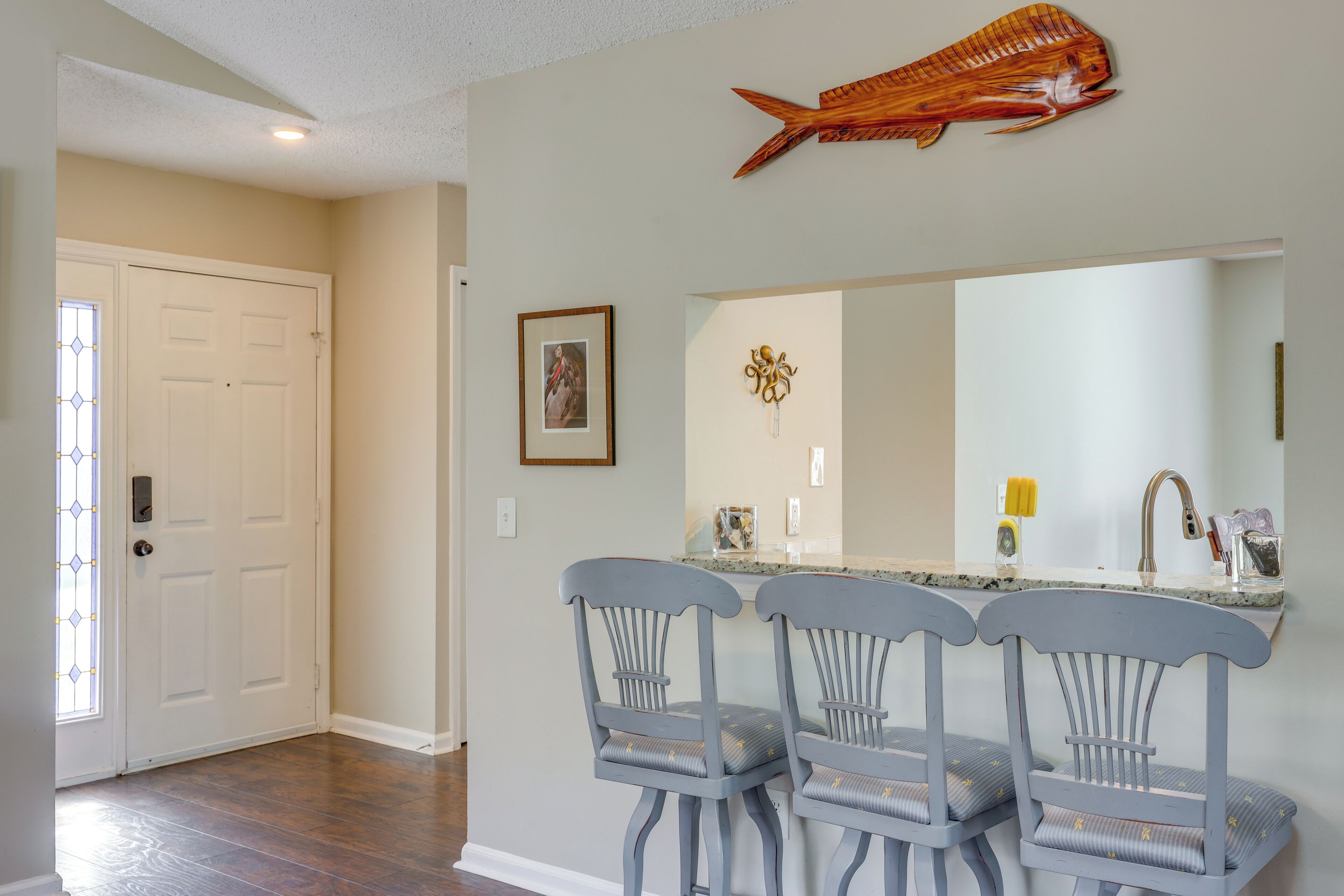 A kitchen with a large island, two bar stools, a wooden fish decoration, a framed picture, and a door with a stained glass window.