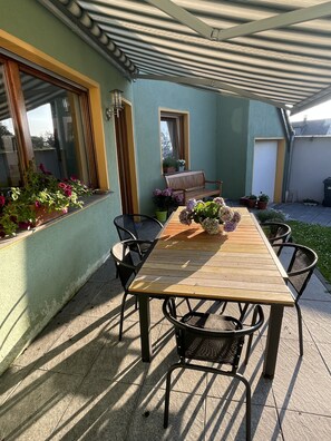 Um pátio externo com mesa e cadeiras de madeira, cercado por plantas em vasos e um banco.