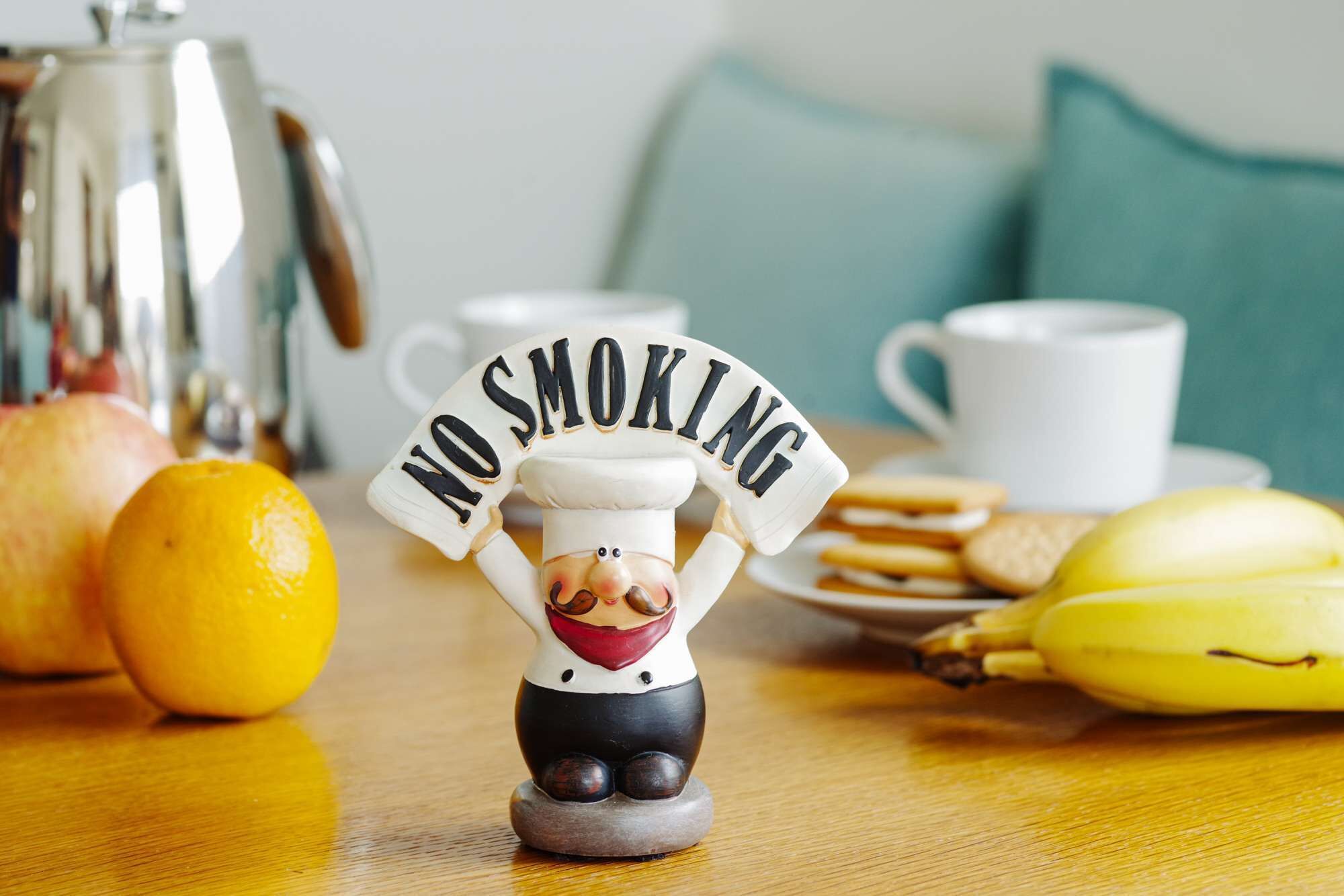 A figurine of a chef with a 'NO SMOKING' sign, surrounded by fruits and a coffee pot on a table.