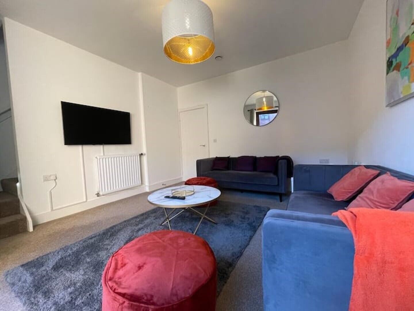 Un salon avec un canapé gris, un pouf rouge et une table basse ronde. Un téléviseur à écran plat est fixé au mur, et un miroir est accroché au-dessus du canapé.