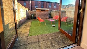 Un pequeño patio trasero con césped artificial, una casita de juegos de madera y cuatro sillas de plástico rojas.