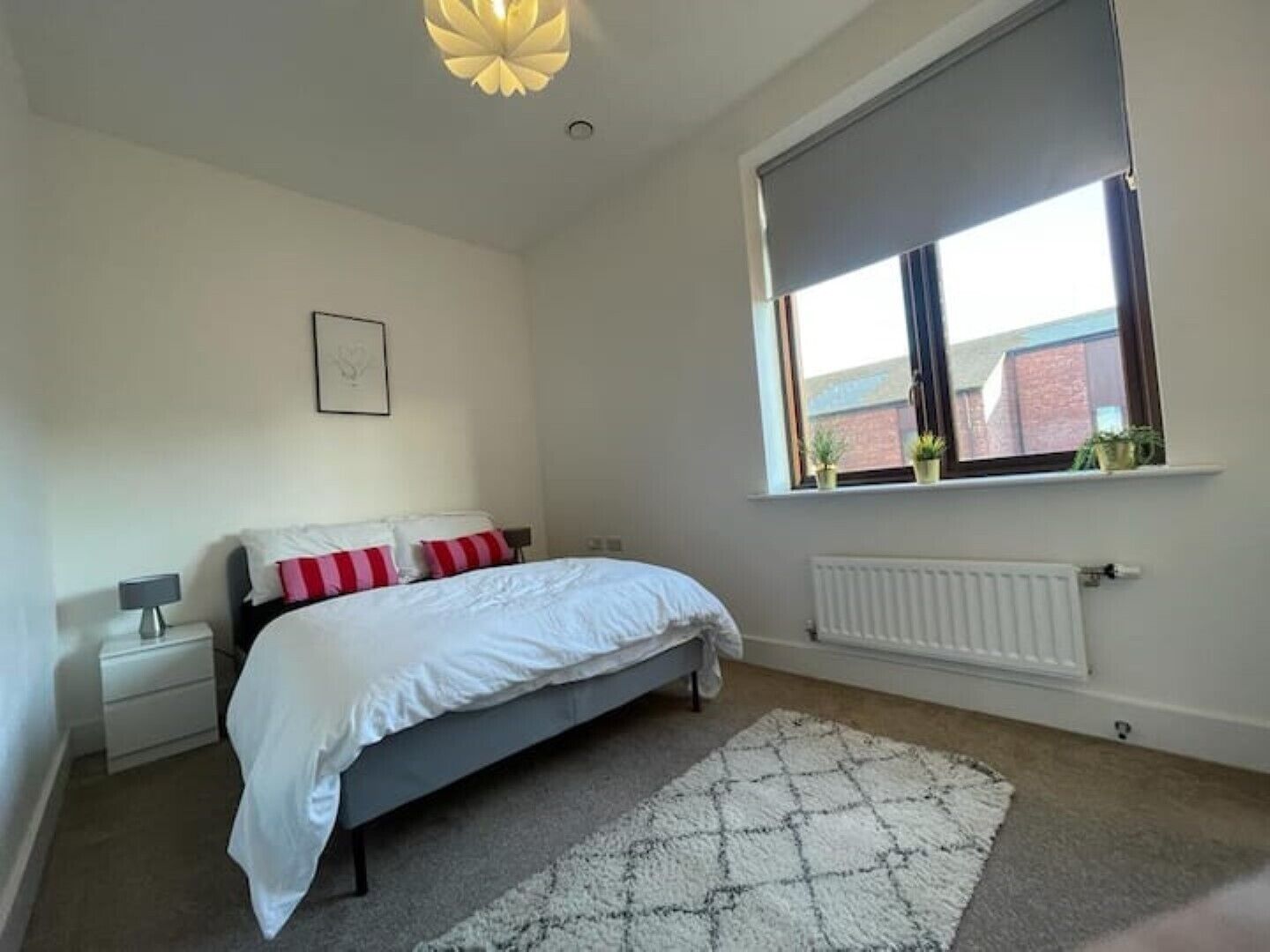 A bedroom with a bed, bedside table, lamp, radiator, window with plants, and a ceiling light fixture.
