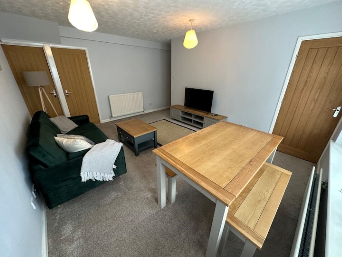 A compact living space with a green sofa, a wooden dining table, a TV, and a radiator.