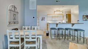 Un comedor con mesa de madera y sillas blancas, una cocina con microondas y taburetes de bar, y una pared azul decorativa.