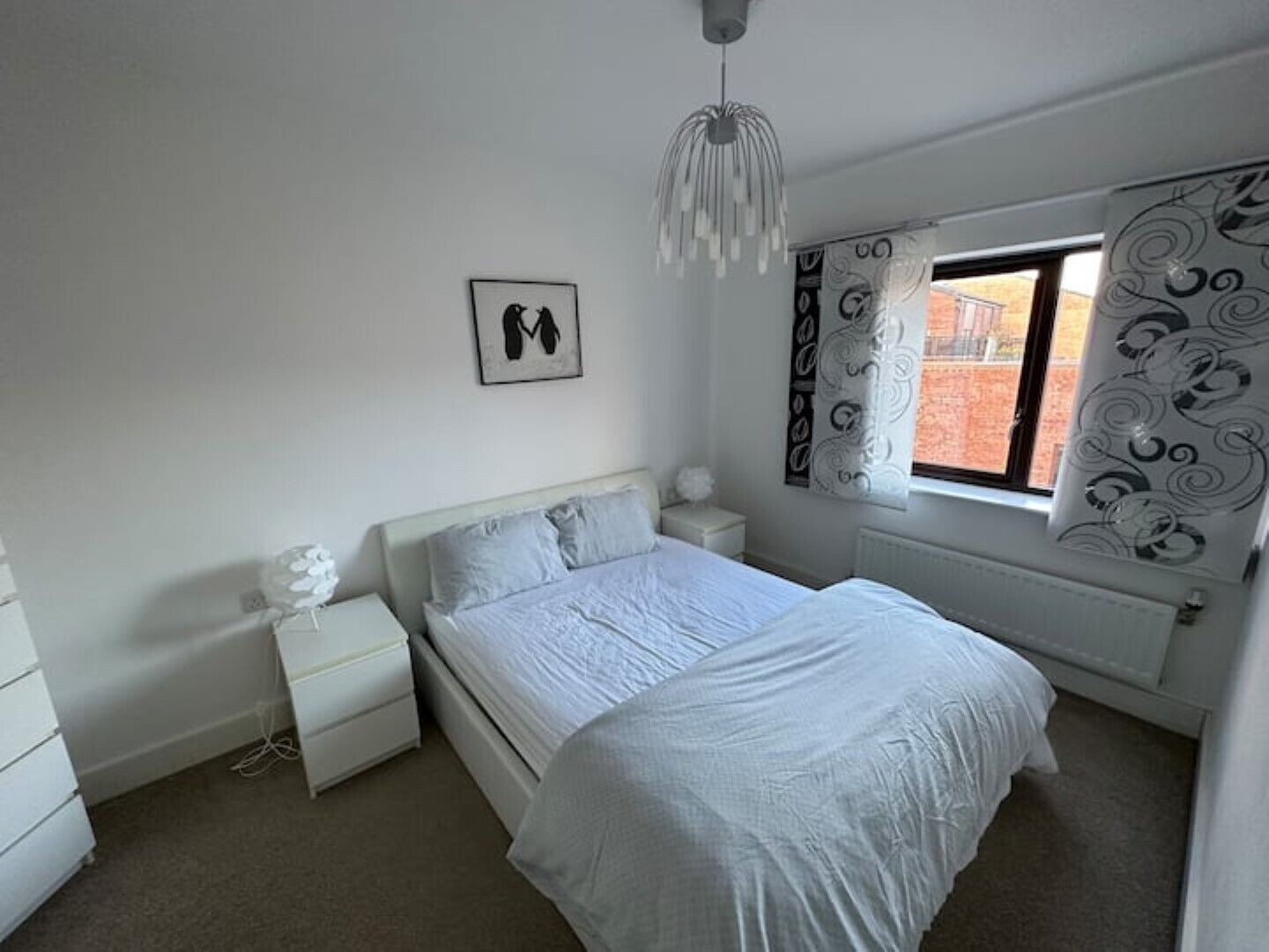 A bedroom with a bed, nightstands, a dresser, a hanging light fixture, and a window with patterned curtains.