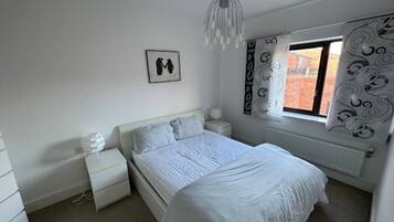 A bedroom with a bed, nightstands, a dresser, a hanging light fixture, and a window with patterned curtains.