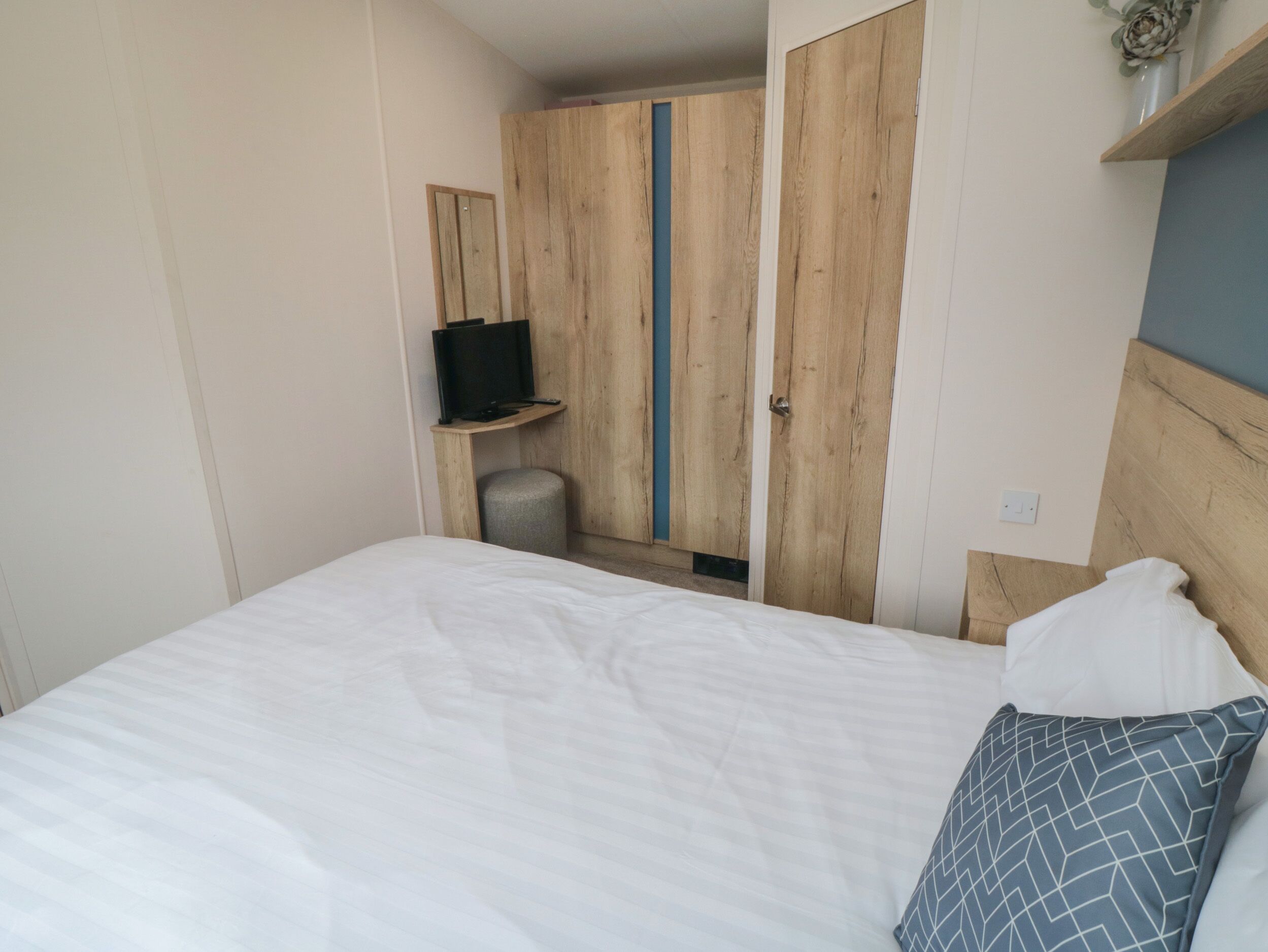 Un dormitorio con cama, televisión sobre un estante de madera, armario y un taburete pequeño.