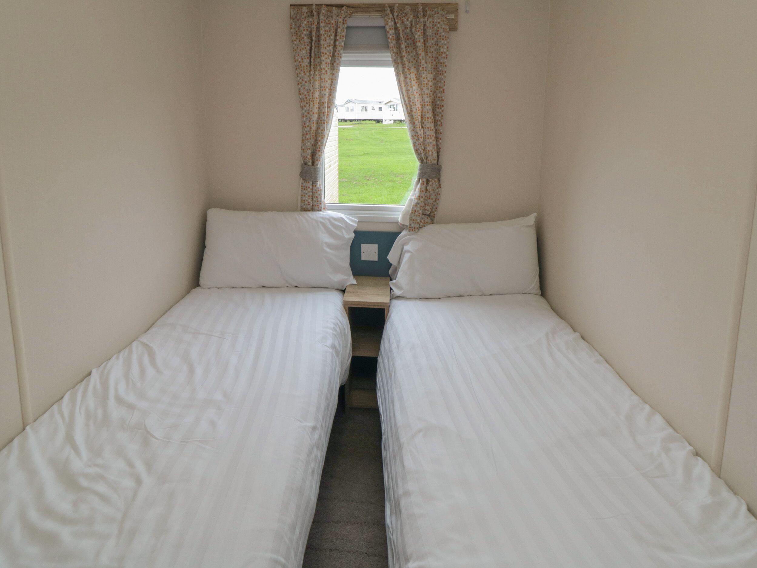 A small, single-room accommodation with two single beds, a window with curtains, and a view of a grassy area outside.