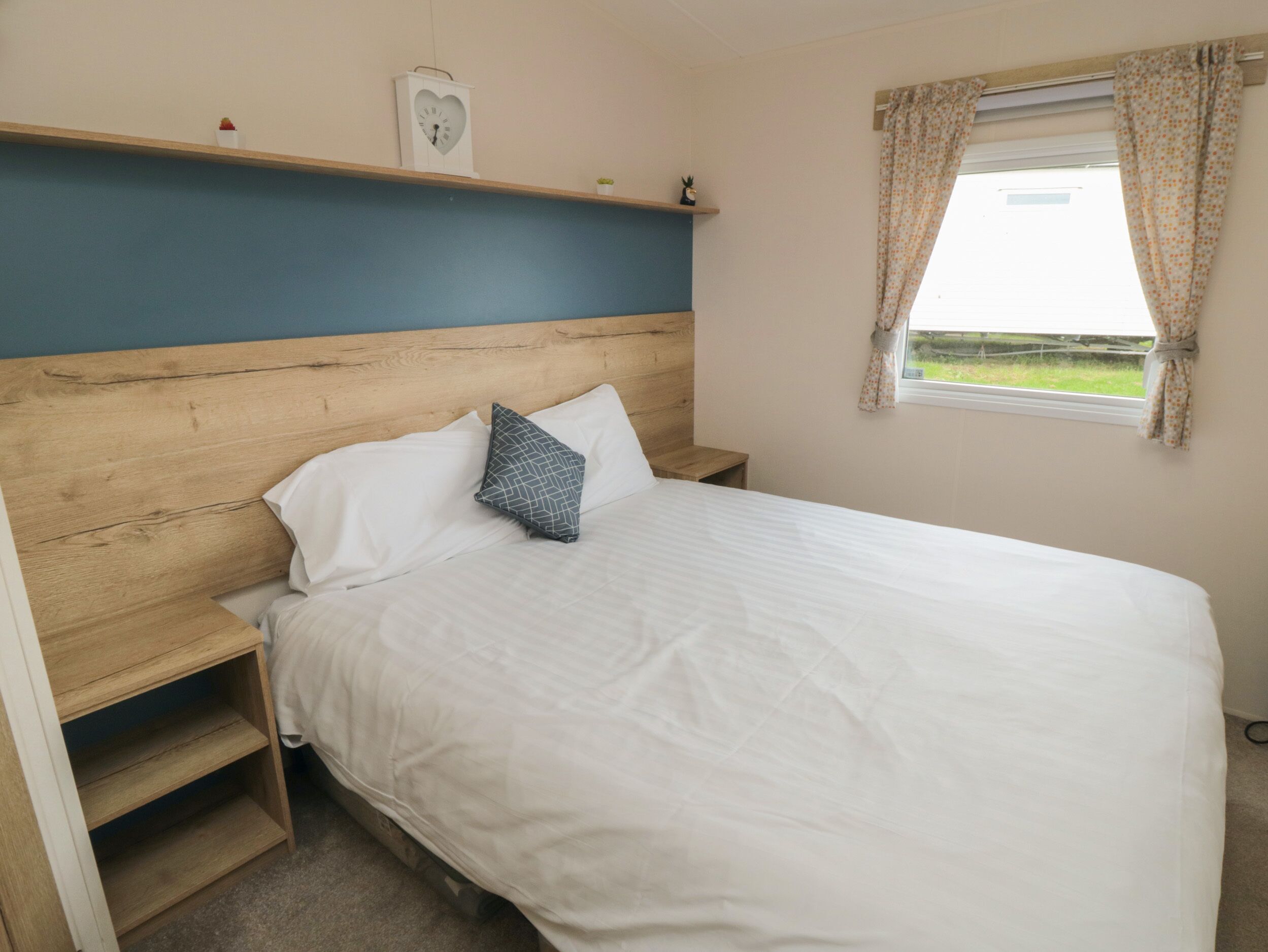 A bedroom with a wooden headboard, a single bed with white bedding, a nightstand, a small shelf with a clock and a plant, and a window with curtains.
