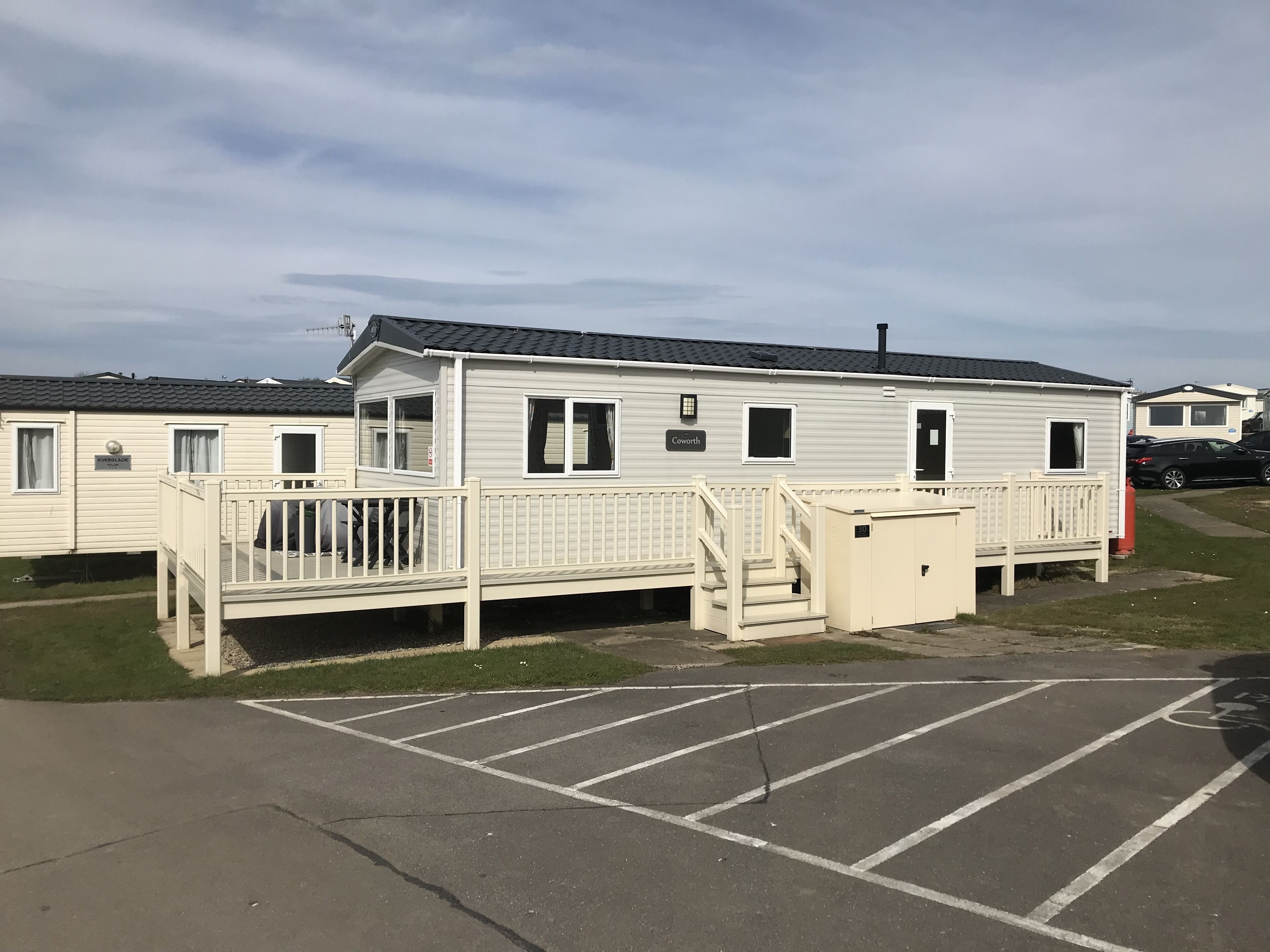 A caravan with a deck and a small attached structure, possibly a utility room, in a caravan park with marked parking spaces.