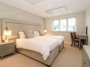 A bedroom with a large bed, bedside tables, a desk with a mirror, and a window with blinds. - The Birches (Windermere)