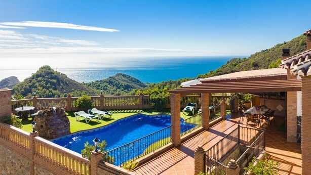 A pool with a view of the sea, a patio with outdoor seating, and a mountain backdrop.