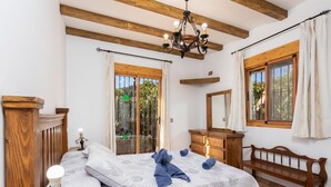 A bedroom with a bed, wooden headboard, a dresser, a bench, a chandelier, and large windows with curtains.