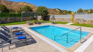 Une piscine rectangulaire bordée de pierres, entourée de chaises longues et agrémentée d’un fauteuil suspendu, le tout adossé à une colline.