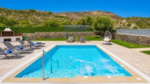 A rectangular swimming pool with clear blue water, surrounded by lounge chairs and a hanging chair, with a stone wall and greenery in the background.