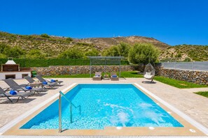 Ein rechteckiger Swimmingpool mit klarem, blauem Wasser, umgeben von Liegestühlen und einer Hängematte, mit einer Steinmauer und Grünflächen im Hintergrund.