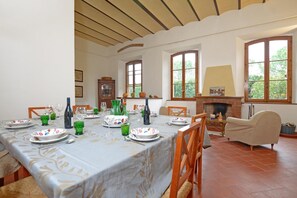 Comedor con una mesa dispuesta para una comida, rodeada de sillas, una chimenea y amplios ventanales.