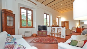A spacious living room with white sofas, a television, and a kitchen area with wooden cabinets and a dining table. - Villa Piandisette - Four Bedroom Villa, Sleeps 10 (Cetona)