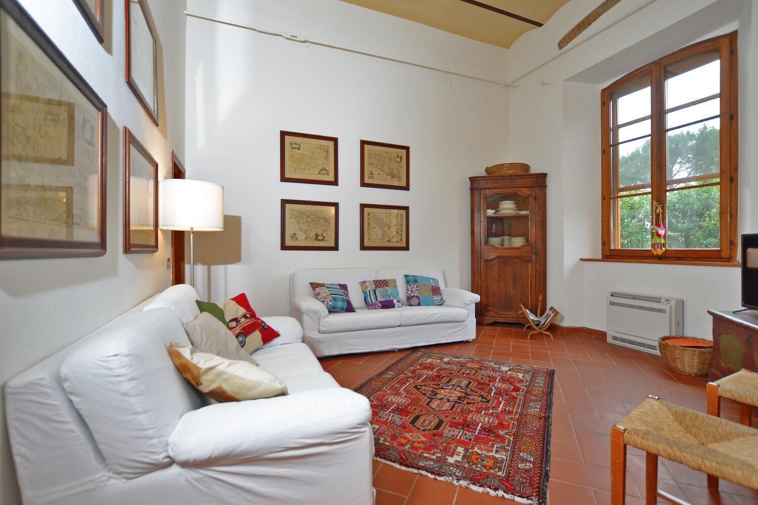 Ein Wohnzimmer mit einem weißen Sofa, einem gemusterten Teppich, einer Holzkommode und einem großen Fenster mit Blick auf Bäume.