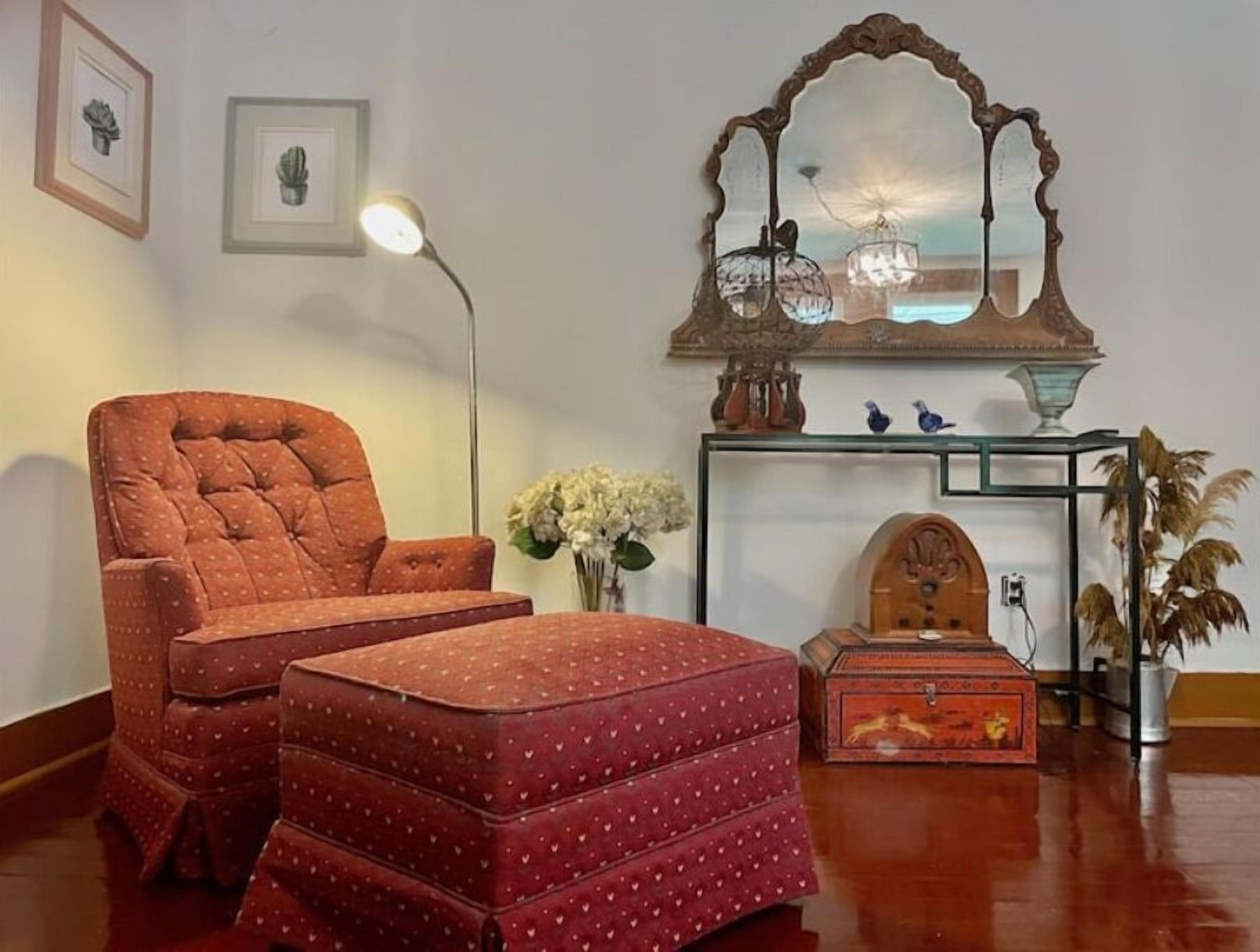 A cozy living room with a tufted armchair, a matching ottoman, a floor lamp, a decorative mirror, and a small side table with a vase of flowers.