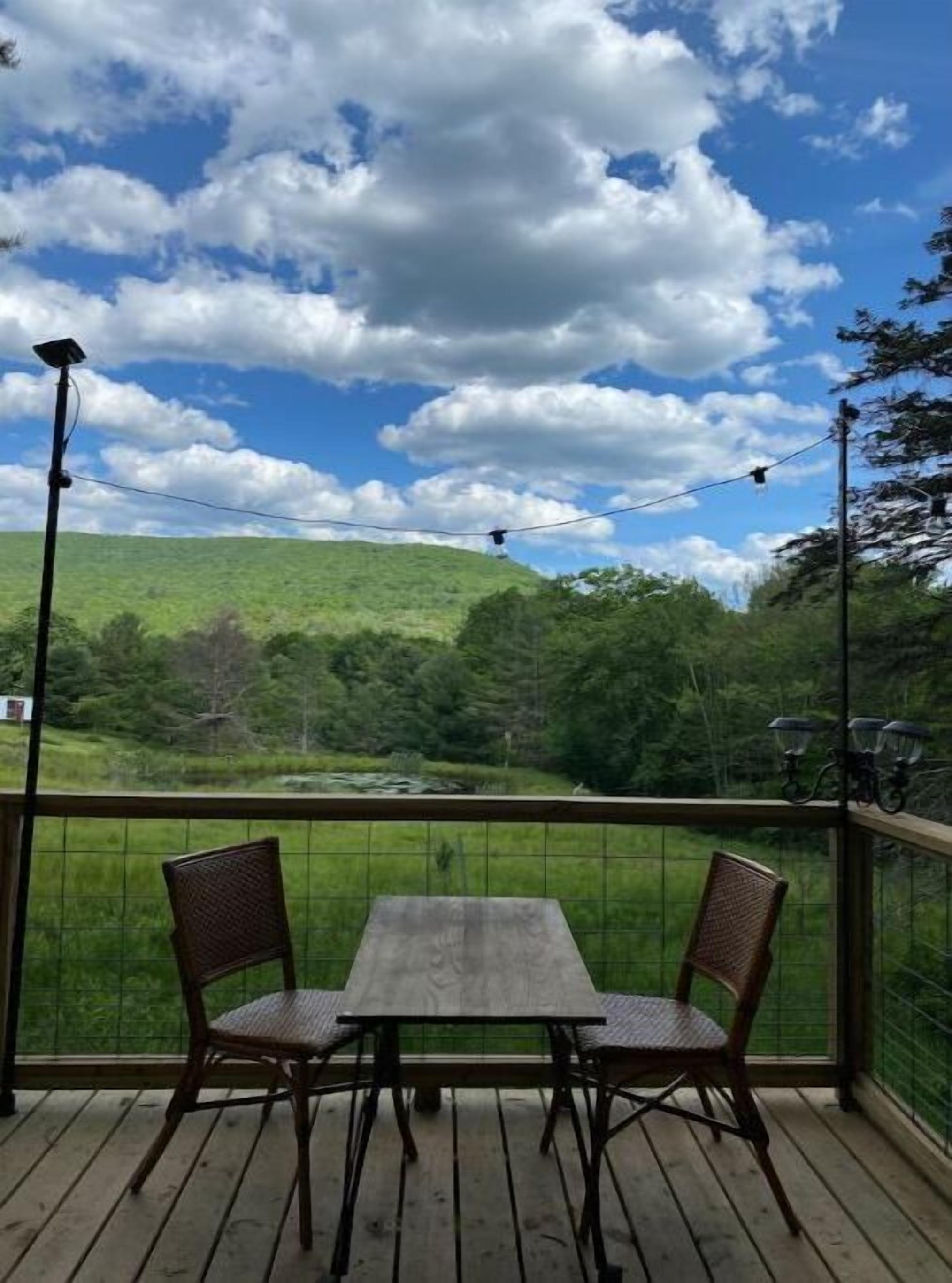 A wooden deck with a table and two chairs, overlooking a green landscape with hills and trees.