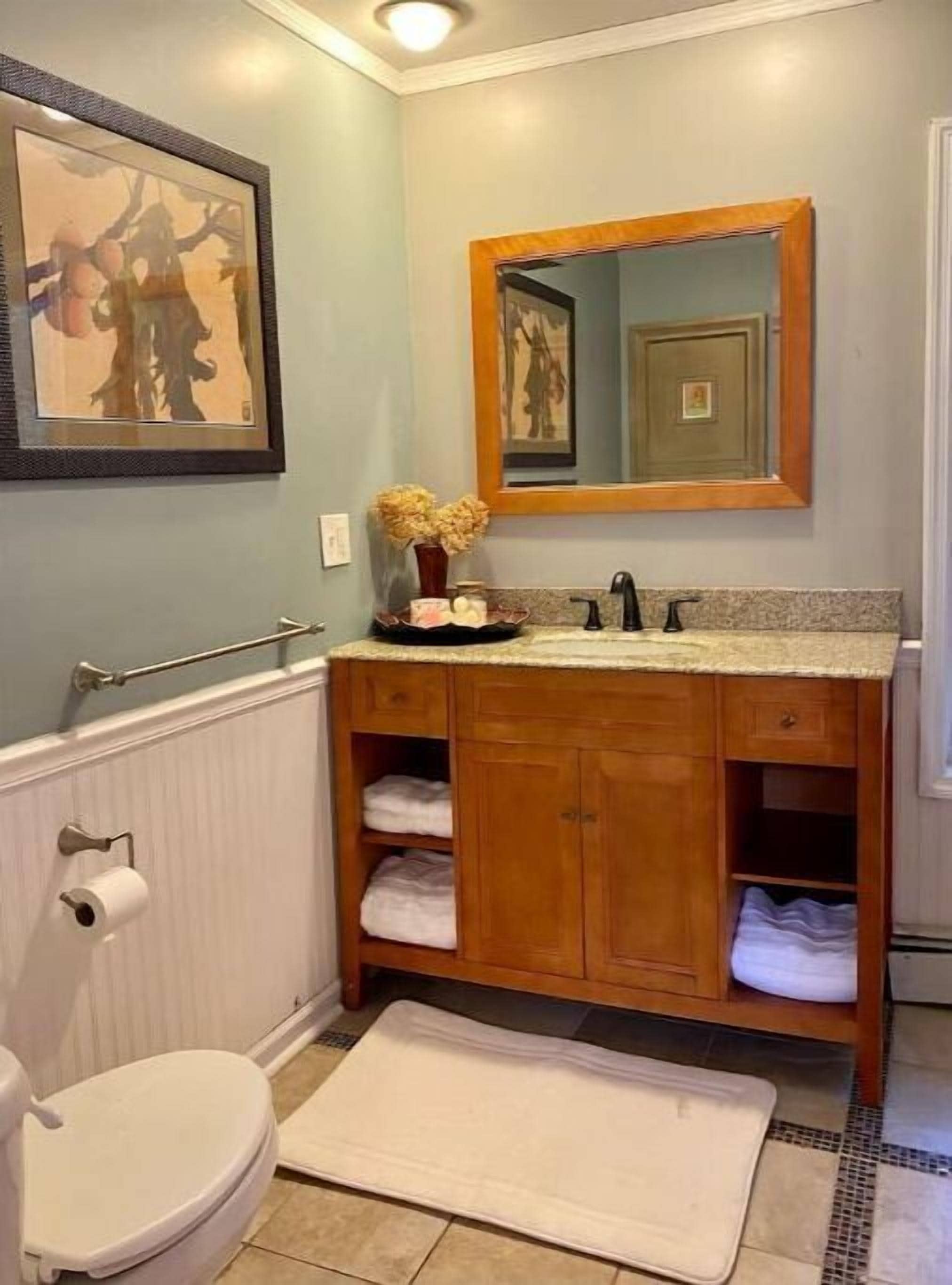 Un bagno con un mobile vanitario in legno, uno specchio, un quadro incorniciato, un water e un piccolo tappeto.