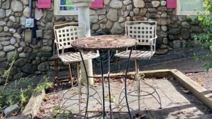 Een stenen gebouw met roze luiken, een kleine tuin met een tafel en stoelen, en een stenen schoorsteen.