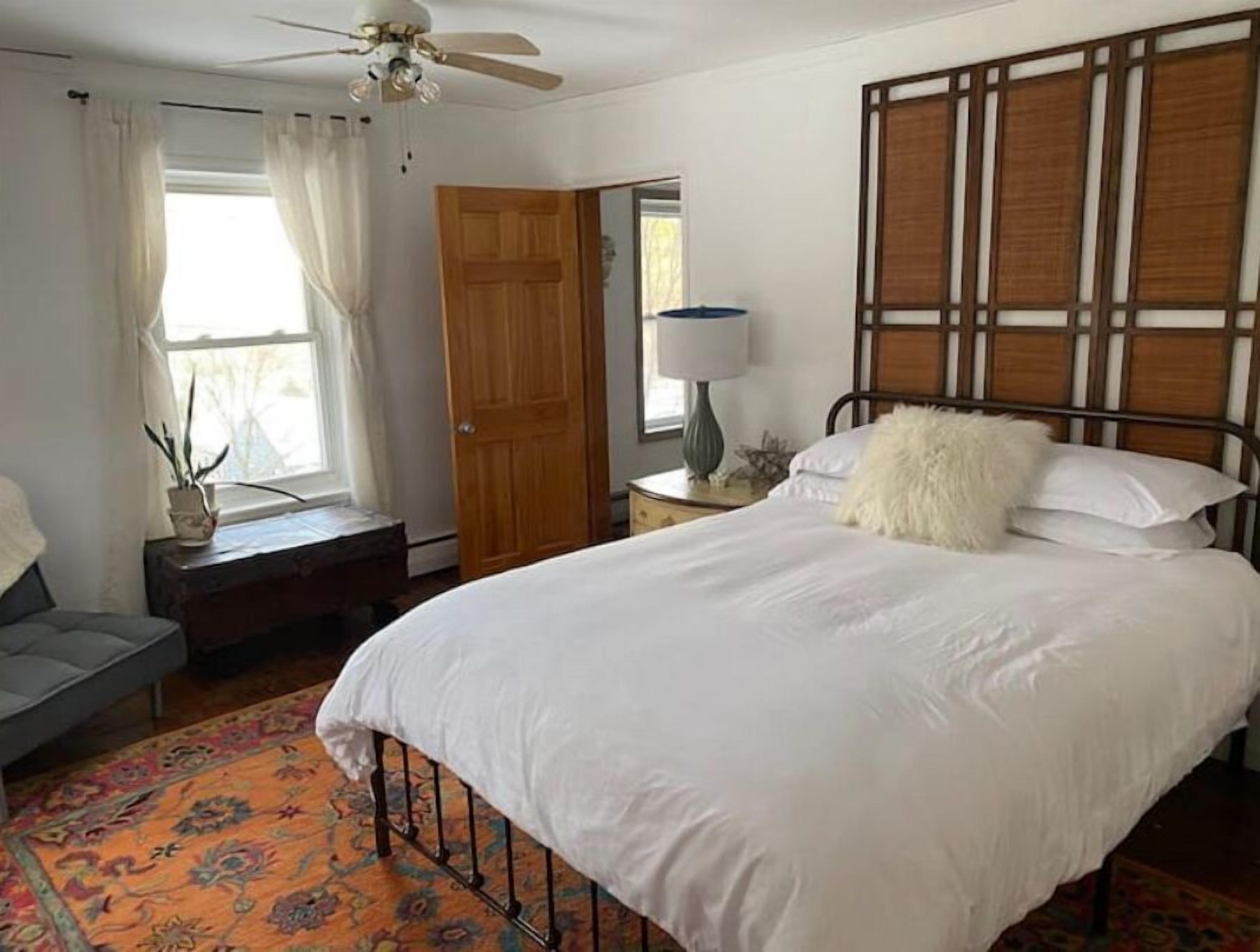 Ein Schlafzimmer mit Bett, Deckenventilator, Fenster mit Vorhängen, Holztür, Nachttisch mit Lampe und dekorativer Kopfteil.