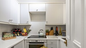 Ett kompakt kök med vita skåp, en spis, en ugn och en bänkskiva med diverse föremål, som en vattenkokare, bananer och ett vinställ.