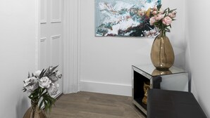 En hall med en vit dörr, en tavla på väggen och två vaser med blommor på ett avlastningsbord.