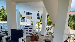 A covered outdoor seating area with white furniture and blue cushions, surrounded by potted plants and a stone floor.