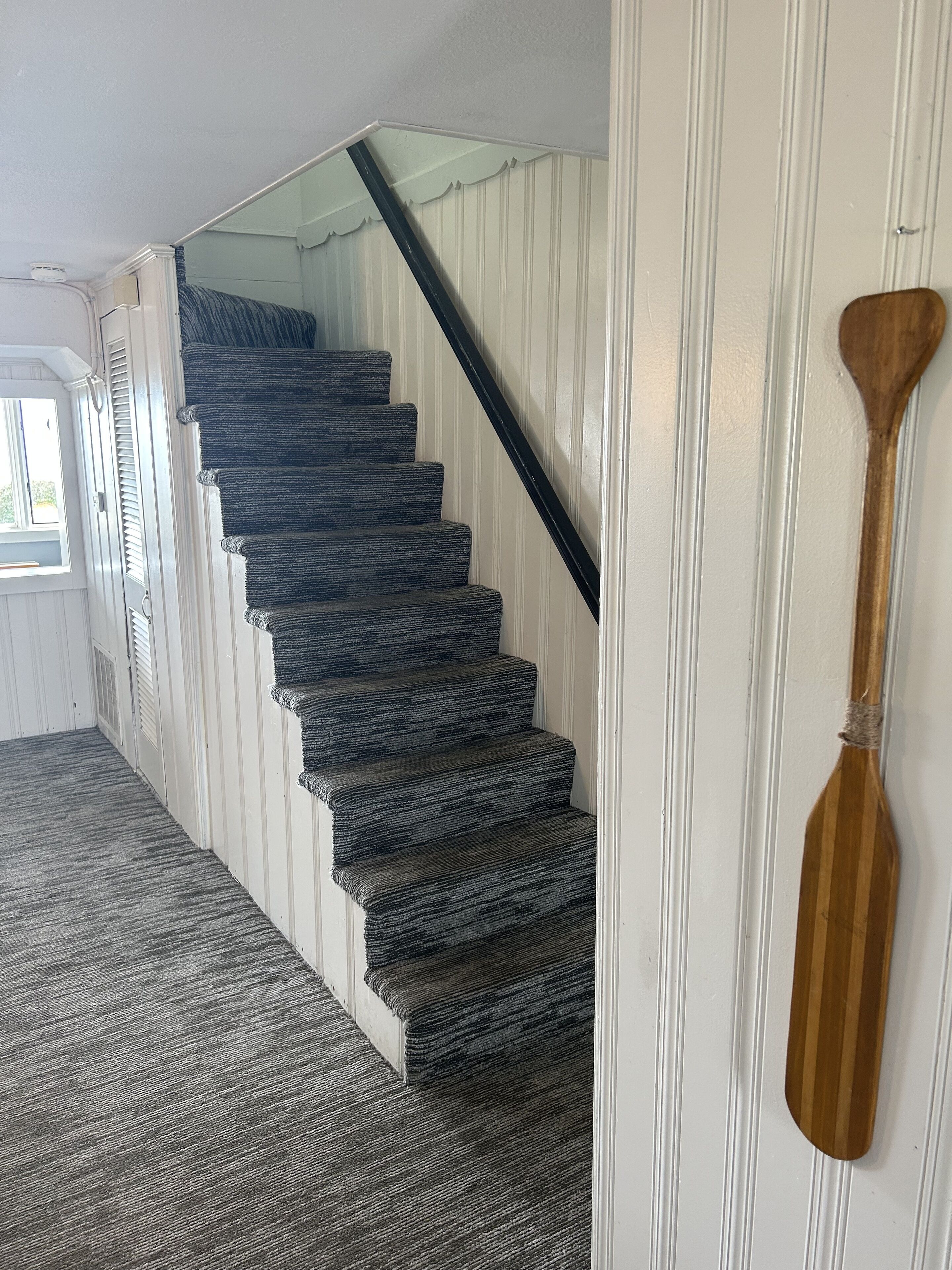 Un escalier recouvert d’une moquette à motifs sombres, avec une cuillère en bois appuyée contre le mur.