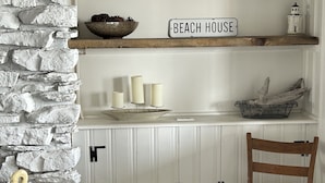 A room with a stone wall, a wooden chair, a small table, and a shelf with a 'BEACH HOUSE' sign. - Lakeside Oasis - Lake Erie Lakefront with Golf Cart (Luna Pier)