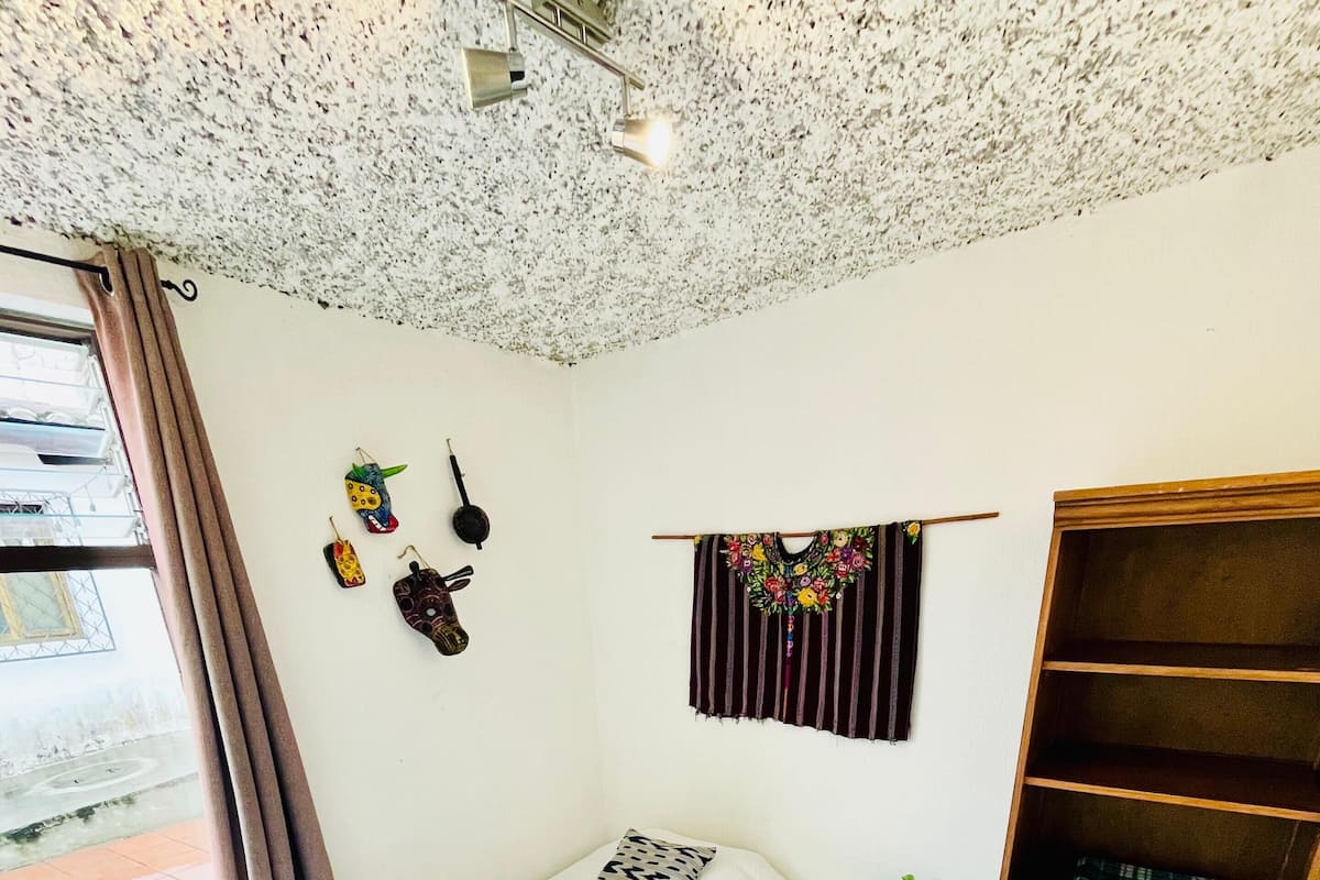Una habitación pequeña y ordenada con una cama, un armario de madera, una mesita y una ventana con cortinas.