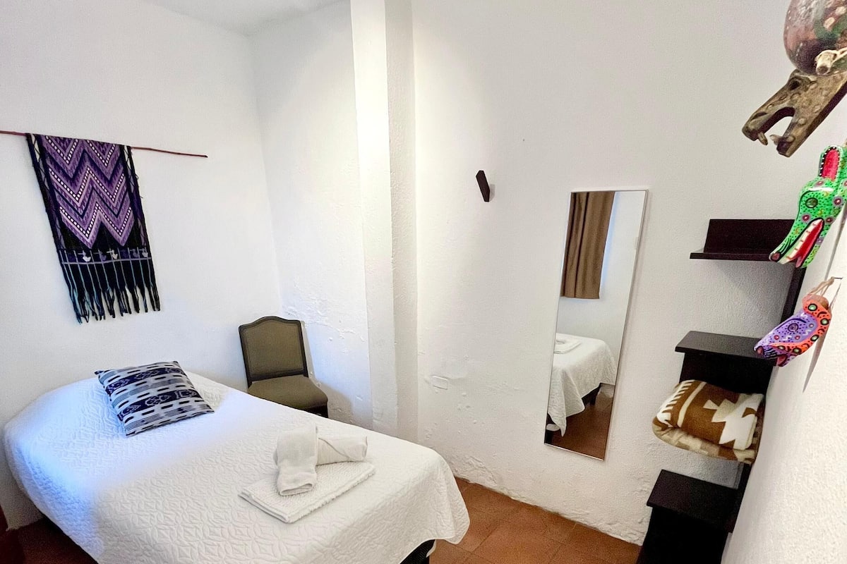 Una habitación pequeña y limpia con cama, una silla, un espejo y un estante con adornos.