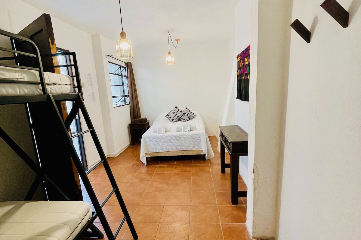 Una habitación compacta con una litera, una cama individual, una mesita pequeña y una lámpara colgante.
