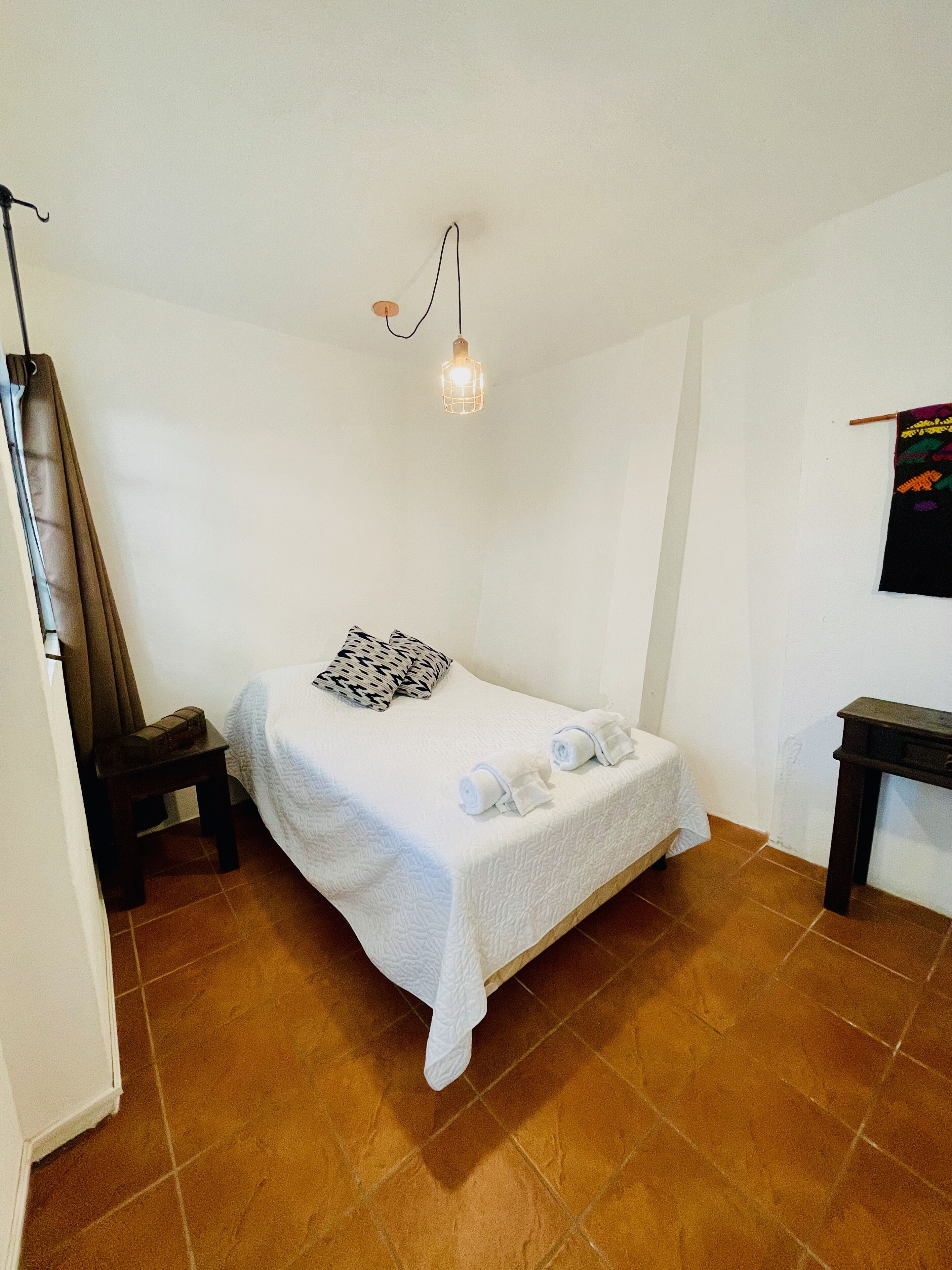 A small, neatly made bed with a white bedspread and two rolled towels, a single black and white patterned pillow, a small wooden nightstand, a wall-mounted light fixture, and a small table with a black cloth.
